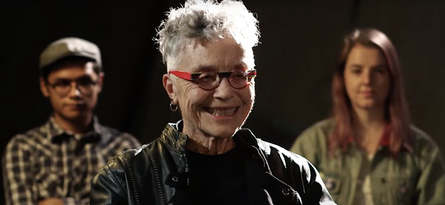 Barbara Hammer, wearing a leather jacket and red framed glasses, stands between two other individuals and smiles toward camera